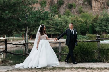 Düğün kıyafetli bir çift, bir su kütlesinin yanında yürür. Sahnede arka planda yeşil ve engebeli kayalar yer alıyor, açık havada bir düğün kutlaması sergileniyor..