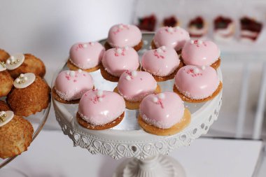 A group of heart-shaped pink desserts sits on a decorative stand at a dessert table. Guests chat and eat in the background at the lively event celebrating togetherness and joy.