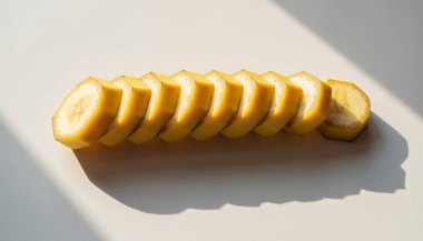 Dilimlere ayrılmış taze bir muzun yakın plan görüntüsü, düz beyaz arka planda düzgün bir sıra halinde dizilmiş. Parlak, doğrudan güneş ışığı farklı bir gölge yaratır..