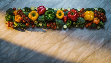 Renkli biber ve sebzeler, yukarıdan çekilmiş hafif desenli bir yüzeyde uzun bir sıra halinde sıralanmış.