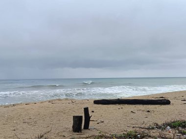 Karanlık fırtına bulutlarının altında sakin bir sahil, dalgalı okyanus dalgaları kumlu kıyılarla buluşuyor. Dalgalar ve kırık direkler kumsalda dinleniyor, sağanak yağmurdan önce karamsar bir kıyı manzarası yaratıyor..