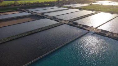 Havadan bakıldığında, sayısız dikdörtgen tankı olan bir su arıtma tesisi görülüyor. Tanklar suyu kahverengi ve mavinin değişik tonlarında tutarlar. Güneş ışığı suyun yüzeyinden yansıyor..