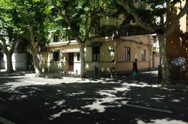 Yapraklı ağaçların altındaki binanın önünden geçen kişi.