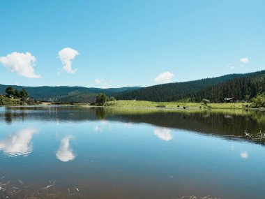 Sakin göl mavi gökyüzünü ve bulutları yansıtıyor.
