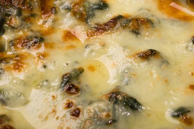 Erimiş peynirle kaplı fırınlanmış ıspanağın yakın plan görüntüsü, hafif yanmış, altın-kahverengi noktaları ve parlak, yağlı bir yüzeyi gösteriyor, zengin dokuyu vurguluyor ve konforlu yiyecekleri cezbediyor..