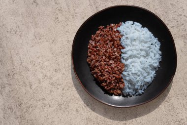 Black Bowl 'da iki çeşit pirinç, Top Down View. Beyaz ve kahverengi pirincin parlak, yüksek açılı görüntüsü koyu bir kaseye bölünmüş. Doğal ışık, nötr arka plan, temiz estetik. Odaklanma keskindir.