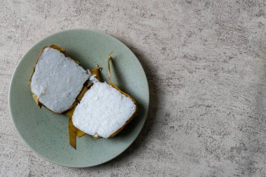 Teal Tabağı 'nda iki parça buğulanmış pirinç keki. Muz yapraklarına sarılı iki beyaz pirinç kekinin yukarıdan çekimleri sessiz bir deniz mavisi tabağında sunuluyor. Yumuşak, doğal ışık ve nötr b.