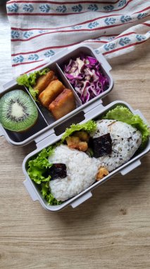 Bento Kutusu Öğle Yemeği: Pirinç, Kivi, Tavuk ve Lahana. Beyaz pilav, kivi dilimleri, kızarmış tavuk ve mor lahana ile bölümlendirilmiş bento kutusunun üstünden. Parlak, doğal ışıklandırma. C.