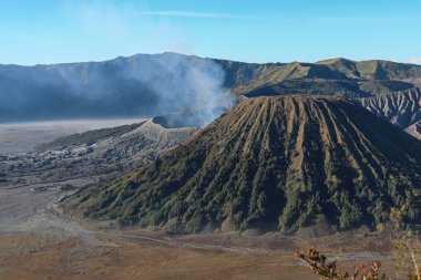 Bromo Volkanik Patlama Dağı, Java, Endonezya. Kraterden yükselen dumanla geniş bir volkanik manzara görüntüsü. Sıcak, dünyevi tonlar açık mavi bir gökyüzü altında sahneye hakim. Dramatik Lig.