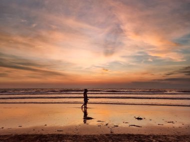 Sunset 'te Sahilde Yürüyen Kişinin Silueti. Sıcak, altın saat ışıklandırması. Islak kumsalda yürüyen bir kişinin silueti. Turuncu ve pembe bulutlu dramatik bir gökyüzü. Sakin okyanus dalgası.