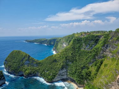 Kelingking Beach Cliff View, Nusa Penida. Turkuaz bir körfez ve beyaz kum plajına bakan yemyeşil bir sahil uçurumunun dramatik hava manzarası. Parlak gün ışığı, berrak mavi gökyüzü dağınık.