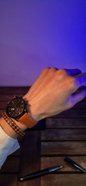 Bulanık neon ve mavi ışıklara karşı bir adamın bileğindeki özel saatin yakın çekimi. Reklamlar için kopyalama alanı olan sanatsal biçim.