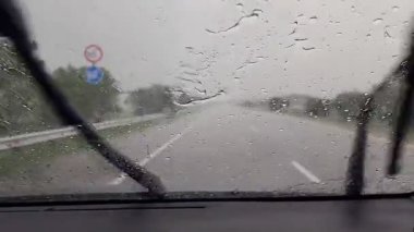 Araba ön camındaki yağmur damlalarının giderilmesi araba tenha bir yolda giderken araba silecekleri tarafından temizleniyor.