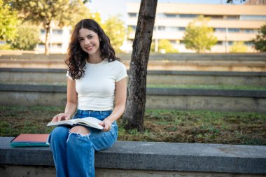 Happy student reading a book while sitting on a bench on university campus