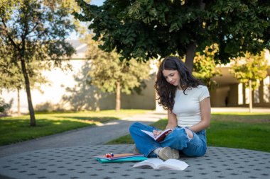 Genç kız öğrenci, üniversite kampüsündeki bir bankta bacak bacak bacak üstüne atarak not defterine notlar yazıyor.