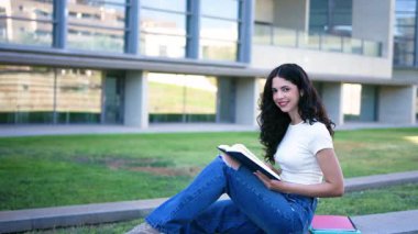 Üniversitede çimlerin üzerinde otururken bir kitapla okuyup ders çalışan genç bir kız öğrenci.