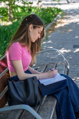 Konsantre olan genç bir kadın, not defterindeki bir kalemle parktaki bankta notlar yazıyor.