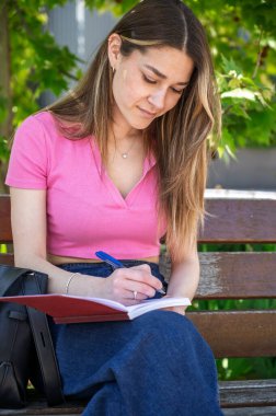 Konsantre olan genç bir kadın, parktaki bankta kalemle deftere yazıyor.