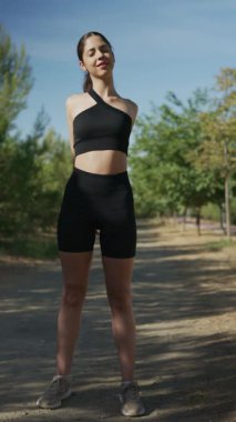 Spor giyimli genç bir kadın halka açık bir parkta ısınıyor ve kollarını açıyor.