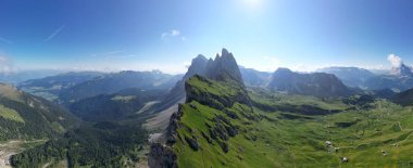 Dolomite dağlarının panoramik hava manzarası keskin tepeleri, yeşil dağlık yamaçları ve açık yaz gökyüzünün altında geniş bir vadi.