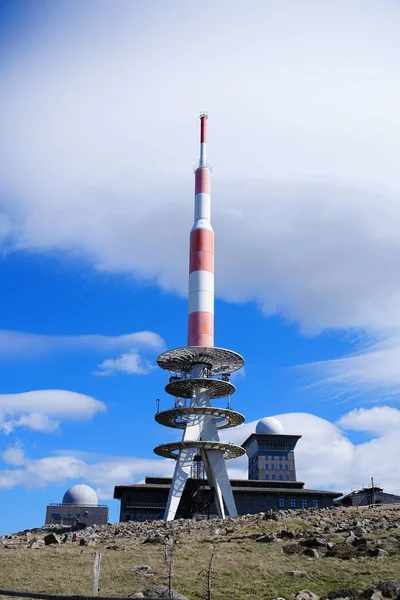 Yüksek bir yayın kulesi, parlak mavi gökyüzü ve dinamik bulutlar altında kayalık bir dağ manzarasının üzerinde yükseliyor.