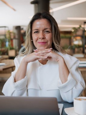 Modern bir kafede çalışan profesyonel bir kadının yakın plan portresi. Ellerini çenesinin altında birleştiriyor ve kurnazca gülümsüyor, güven, güven ve liderlik varlığıyla iletişim kuruyor.
