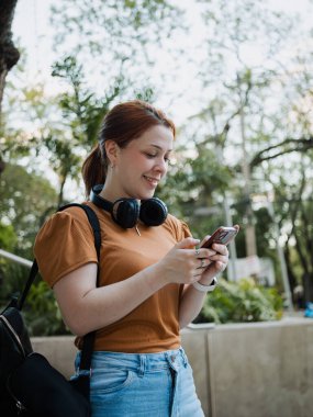 Parkta akıllı telefon kullanan, kulaklık takan, boyun ve sırt çantası takan, cep telefonu mesajlaşması ve dijital yaşam tarzı kavramı olan genç bir kadının yan görüntüsü..