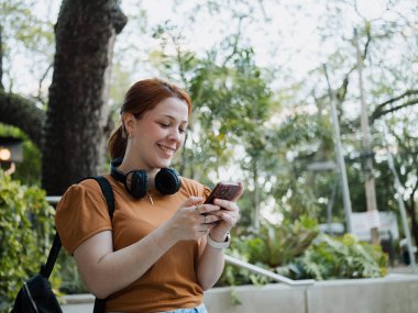 Sırt çantalı ve kulaklıklı genç bir kadın parkta akıllı telefondan mesaj atarken gülümsüyor. Çevrimiçi iletişim ve öğrenci yaşam tarzı kavramı..