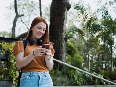 Genç bir kadın açık havada ağaçlar ve yeşillikler eşliğinde akıllı telefon kullanıyor. Kulaklık takıyor, boyun ve sırt çantasına takıyor, gündelik dijital iletişim ve yaşam tarzı kavramı kullanıyor..