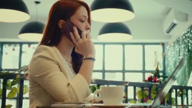 Modern bir kafede dizüstü bilgisayarla çalışırken akıllı telefondan konuşan genç bir iş kadını, kahve fincanı masada, uzaktan çalışma, müşteri desteği ve iş iletişimi konsepti.