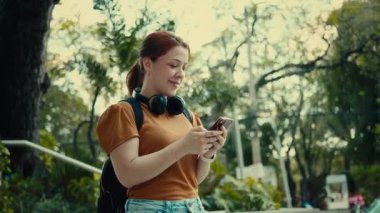 Sırt çantalı ve kulaklıklı gülümseyen genç kadın merdivenlerde akıllı telefon kullanıyor, mesajları mesaj atıyor ve kontrol ediyor, cep telefonu iletişimi ve günışığında modern yaşam tarzı kavramını kullanıyor..