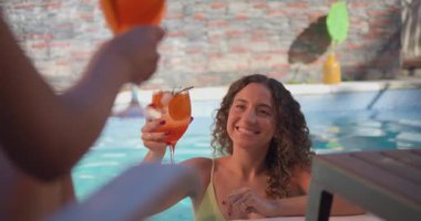 Yüzme havuzunda gülümseyen genç bir kadın kadeh kaldırmak için kadeh kaldırırken bir arkadaş ön planda içkileri tokuşturuyor. Güneşli arka bahçe havuz başında kutlama, yaz yaşam tarzı ve konukseverlik konsepti.