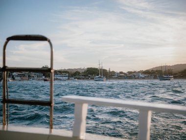 Gün batımında bir tekneden sahil kasabasına ve yelkenlilere doğru bir görüntü, gidilecek yer için hafif dalgalar, yelken yaşam tarzı ve turizm kavramları olan sıcak bir varış sahnesi.