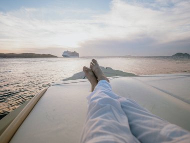 Gün batımında bir teknede rahat ayaklar. Uzakta bir yolcu gemisiyle birlikte. Rahat bir yolculuk anı, yavaş yaşam ve sakin bir okyanus tatili havası..