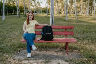 Bir yetişkin, yeşil bir parkta sırt çantasıyla kırmızı bir bankta dinleniyor, sessiz bir bekleme ve düşünme anında duruyor..