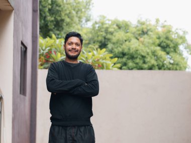 Kollarını kavuşturmuş ve doğal bir gülümsemeyle ayakta duran genç bir adamın portresi, açık havada arka planda bir ışık duvarı ve bitki örtüsüyle. İletişim kurmak için ideal görüntü, modern yaşam tarzı, pozitif tutum, gündelik moda ve reklam mesajları