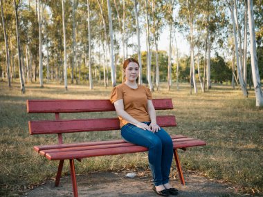 Ormanlık bir parkta kırmızı bir bankta oturan, rahat duruşu ve sakin ifadesiyle genç bir kadının görüntüsü. Yaşam tarzı kavramları için ideal fotoğrafçılık, dinlenme, boş zaman, kentsel doğa, günlük moda kampanyaları veya yerel turlarda refah ve ticari kullanım