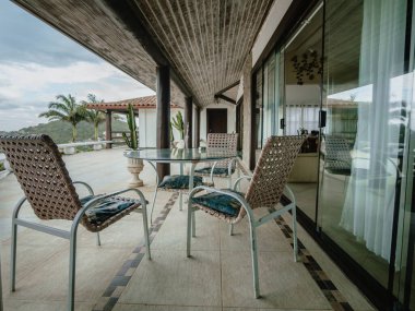 Geniş pencerelerin yanında mobilyaları (masa ve sandalyeleri) olan açık teras, doğal bir çevreye ve konut mimarisine açık bir manzaraya sahip. Konaklama, dış tasarım, yaşam tarzı, turizm ve dinlenme temaları için uygun görüntü.