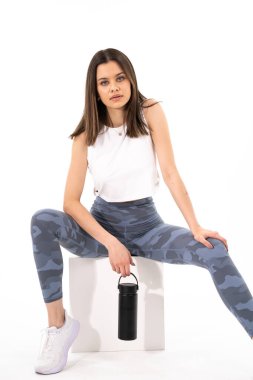 Mavi pantolonlu ve beyaz kolsuz tişörtlü beyaz bir kutuda oturan bir kadın. Elinde siyah bir su şişesi tutuyor.