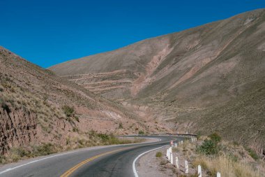 Uzun, kıvrımlı bir yol bir çöl manzarasında esiyor. Yol uzun otlarla kaplı ve görünürde hiç ağaç yok. Gökyüzü açık ve mavidir ve güneş ışıl ışıl parlar.
