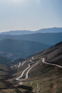 Dağın yamacına çıkan dolambaçlı bir yolu olan bir dağ sırası. Yol ağaçlarla çevrili ve gökyüzü açık ve mavi.
