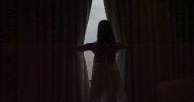 Bir kadın perdesi açık bir şekilde pencerenin önünde duruyor. O, beyaz bir elbise giyiyor ve yalnız. Sahne gizemli ve tüyler ürpertici.