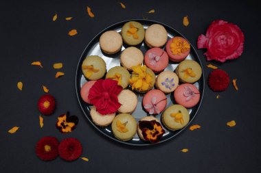 Rengarenk Fransız badem kurabiyelerinin zarif kompozisyonu. Taze bahar çiçekleriyle süslenmiş. Koyu bir arka planda çiçek açan dalları olan bir tabakta. Çiçek süslemeli ve canlı pastel renkleriyle hayat dolu şık bir tatlı..