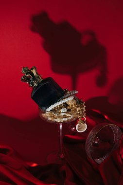 Kırmızı ipek kumaş üzerinde inci ve altın mücevherlerle dolu bir bardağa yerleştirilmiş lüks bir parfüm şişesinin bulunduğu zarif bir yaşam kompozisyonu. Dramatik ışıklandırma ve gölge büyüleyici, lüks bir atmosfer yaratır..