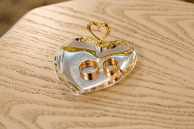 Glass Heart Holder 'da altın alyanslar. Zarif altın alyanslar cam bir kalp tutacağında duruyor. Süslü, aşk evliliği bağlılığını ve kutlamaları simgeliyor.