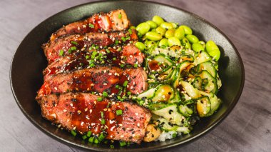 İnce dilimlenmiş sığır filetosu teriyaki, edamame ve sunomono salatası ile güzel bir kase. Yumuşak sığır eti üzerine parlak teriyaki jölesi, taze ve canlı Japon yemeği.