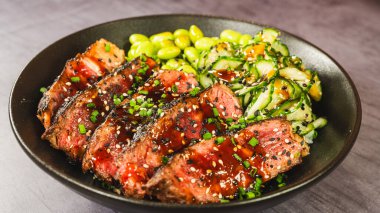 İnce dilimlenmiş sığır filetosu teriyaki, edamame ve sunomono salatası ile güzel bir kase. Yumuşak sığır eti üzerine parlak teriyaki jölesi, taze ve canlı Japon yemeği.