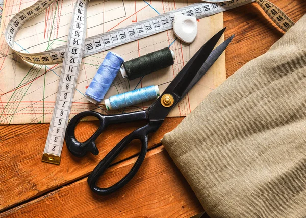 Швейні інструменти, ножиці, шпали ниток, вимірювальна стрічка лежать на діаграмах хвостовиків і дерев'яному столі Стокове Зображення