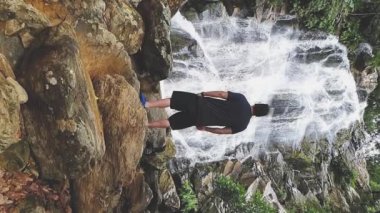 Dikey Video: Adam büyük kayaların üzerinde şelalenin önünde durup suyu seyrediyor, yavaş çekim videosu. Çevre turizmi, macera ya da keşif kavramı. Roque de Minas, MG, Brezilya.