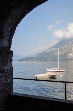 Sabahın erken saatlerinde Como Gölü 'nü taş kemer şeklinde bir pencereden görmek, İtalya, Varenna' ya demirli yelkenli, sisli, yumuşak güneş ışığı ve uzak dağlarla dolu açık bir gökyüzü.